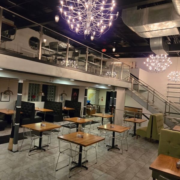 Modern restaurant interior with two levels, chandeliers, wooden tables, and black and green booth seating.