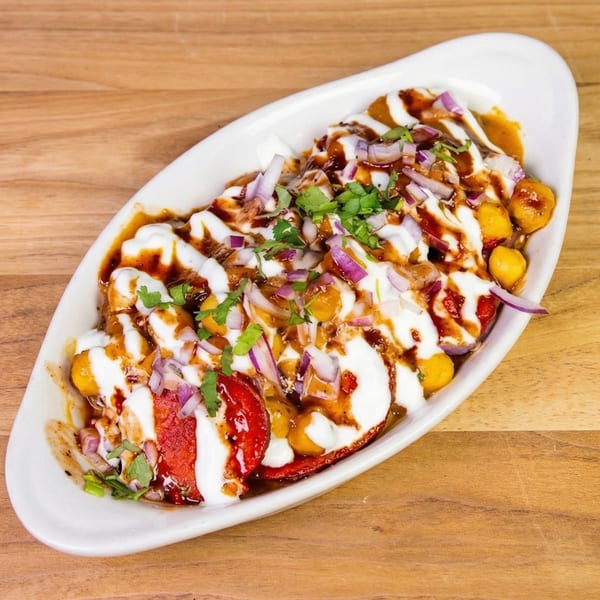 A white oval dish filled with Indian chaat, topped with yogurt, chutney, red onions, and cilantro.