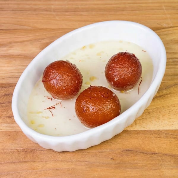 Three gulab jamun, garnished with saffron threads, in a white oval dish on a wooden table.
