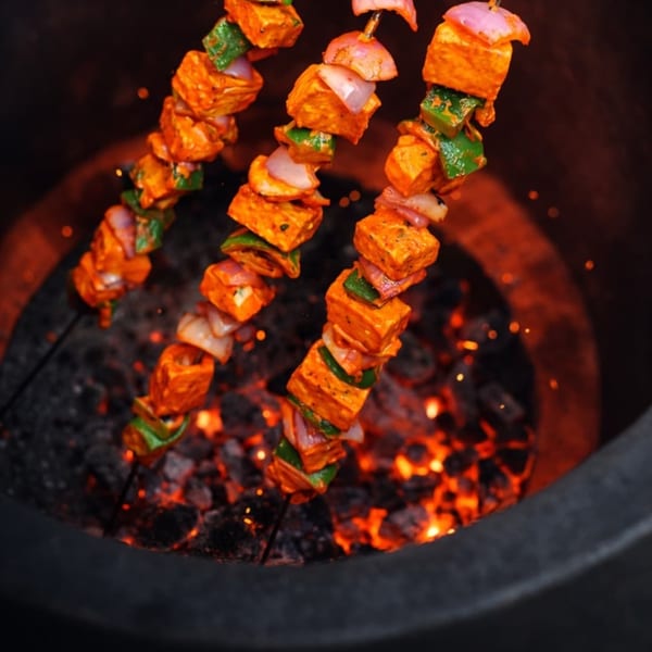 Three skewers of paneer tikka, with cheese, onions, and peppers, cooking over glowing coals.