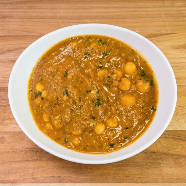 A white bowl filled with a rich, orange-brown stew containing chickpeas and green herbs, on a wooden surface.