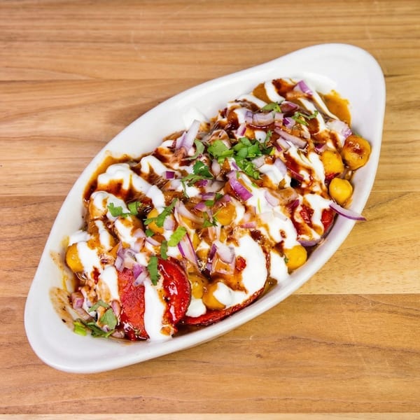 A white oval dish filled with Indian chaat, topped with yogurt, chutneys, red onions, and cilantro.