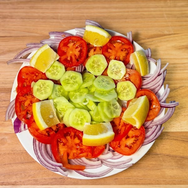 A fresh salad with sliced tomatoes, cucumbers, red onions, and lemon wedges on a white plate.