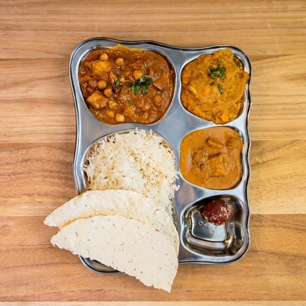 A metal thali plate with rice, papadum, three curries, and a small dessert on a wooden table.