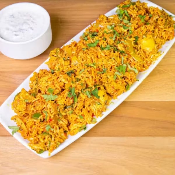 A long white platter of golden biryani with green herbs, next to a small bowl of white sauce.