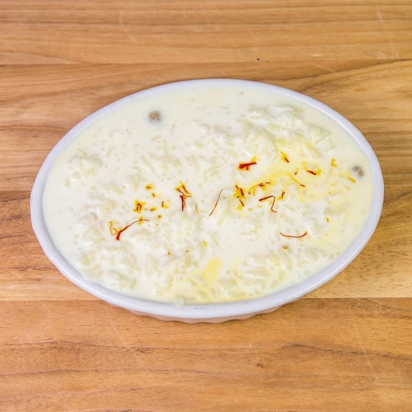 Creamy rice pudding, garnished with saffron threads and nuts, in a white oval dish on a wooden surface.