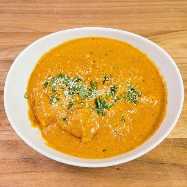 A white bowl of creamy orange curry, garnished with fresh cilantro and grated cheese, on a wooden table.